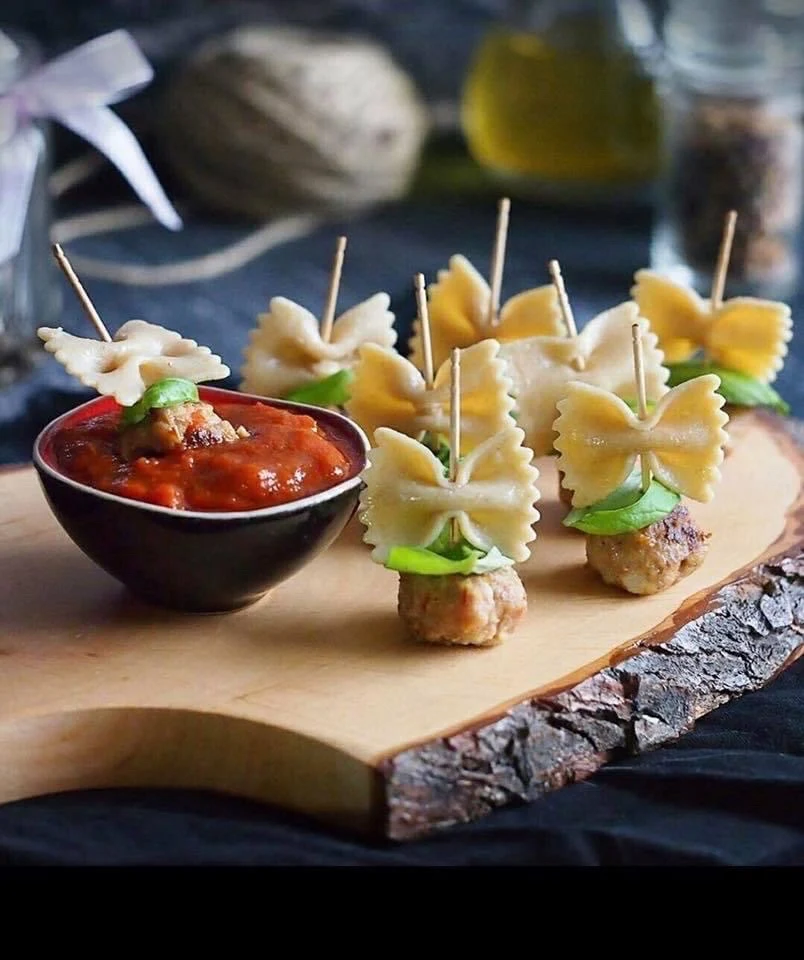 Meatball skewers topped with bow tie pasta and basil leaves, served with a bowl of tomato sauce on a wooden board.