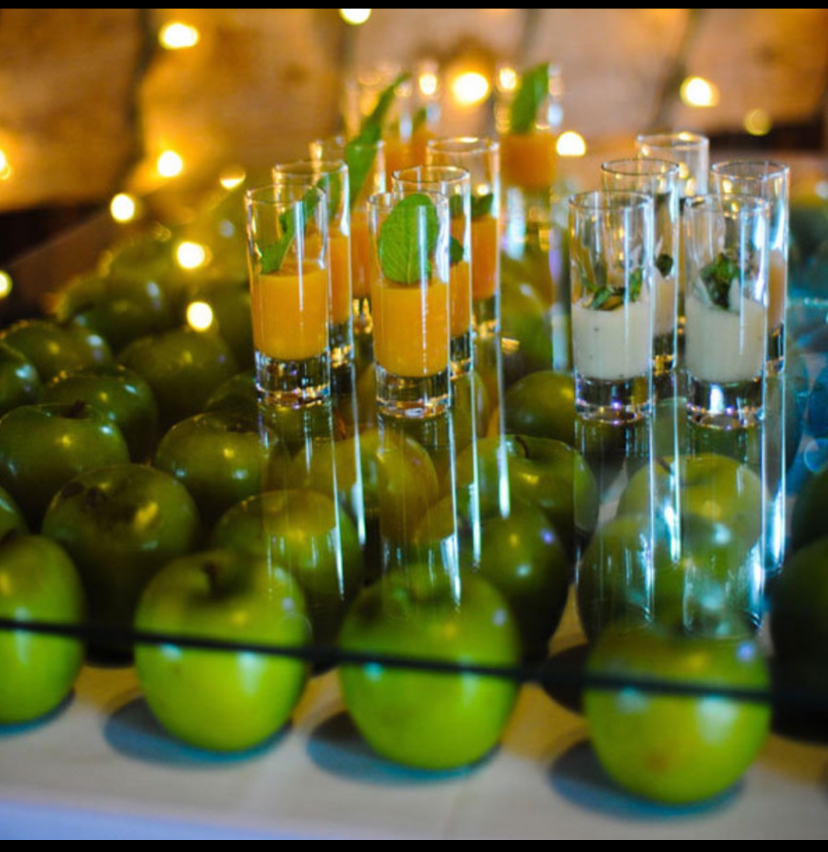 Rows of green apples surround glasses of orange and white drinks, garnished with mint, on a glass-topped table.