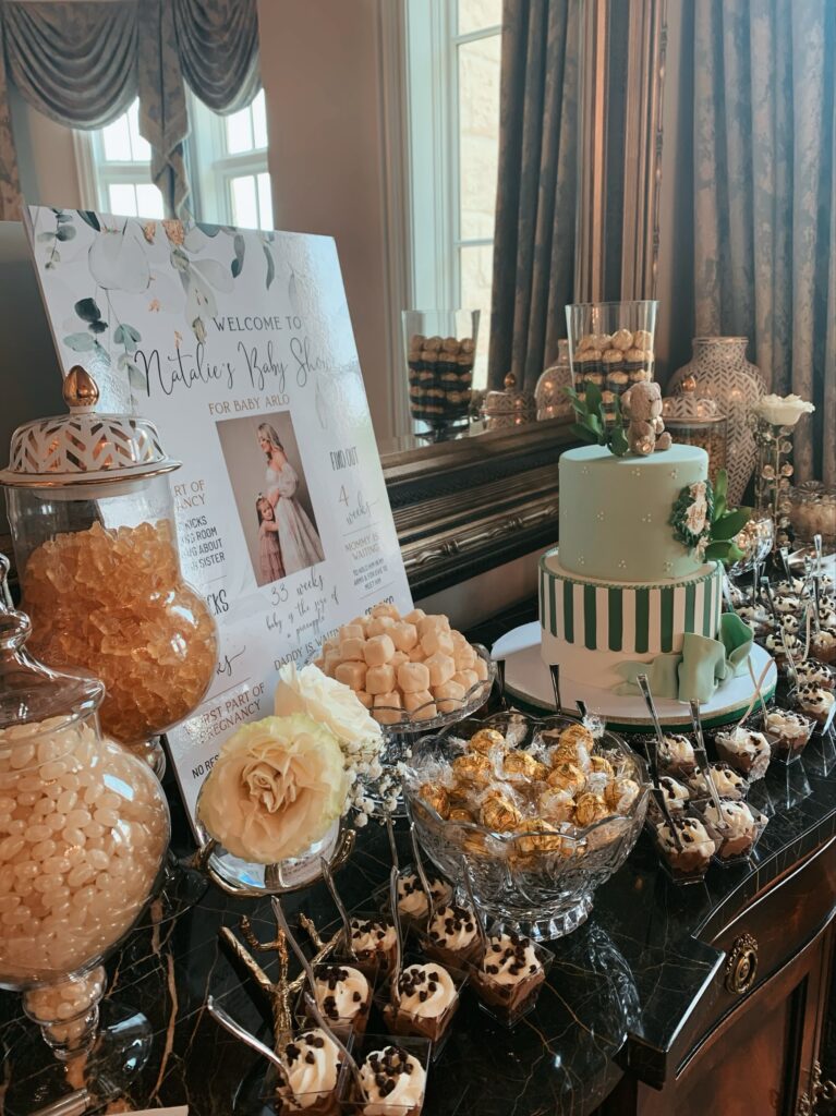 Dessert table with a green cake, candy jars, chocolates, and a baby shower welcome sign displayed by a window.