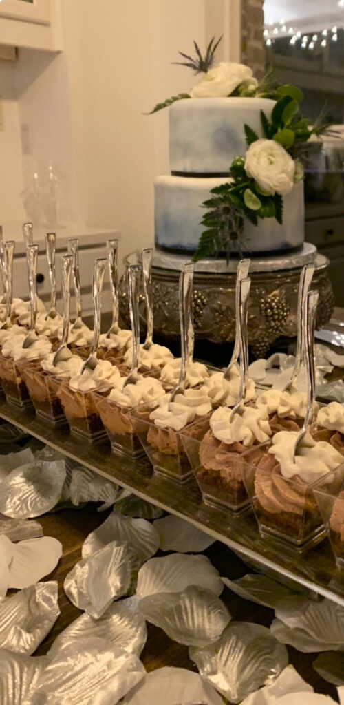 Mini dessert cups with whipped topping are arranged in front of a two-tiered white and blue cake with floral decor.