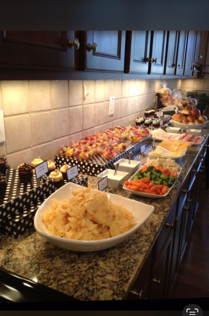 A kitchen counter with chips, cupcakes, fruit skewers, vegetables, cheese, bread, and sandwich fixings arranged for a buffet.