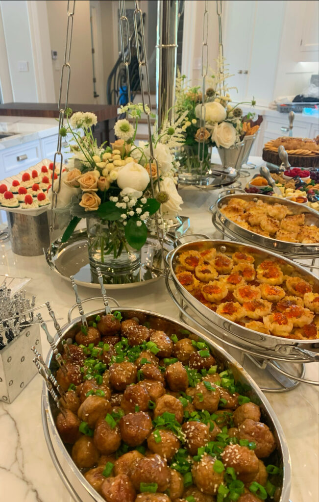 Buffet table with trays of meatballs, pastries, and other appetizers, decorated with fresh white and beige flowers.