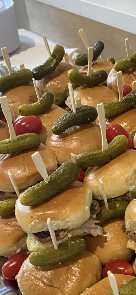 Mini sandwiches topped with pickles and skewered with wooden sticks, garnished with cherry tomatoes on a tray.