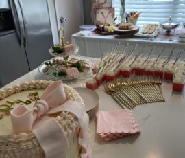 Baby shower dessert table with a decorated cake, snacks, gold forks, and pink napkins arranged on a kitchen counter.