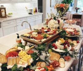 A kitchen island covered with assorted cheeses, fruits, crackers, meats, and flowers, set up for a gathering.