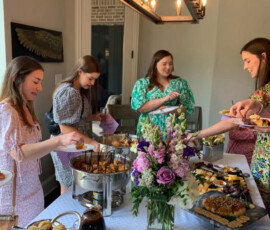 Four women serve themselves food from a buffet table with flowers and assorted dishes in a bright dining room.