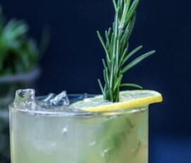 A cocktail with ice, garnished with a lemon slice and rosemary sprig, sits beside a halved lemon on a dark surface.