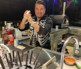 A man in a skeleton shirt shakes a cocktail behind a bar, surrounded by bottles and bar tools.