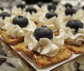 Screenshot Toasted crackers topped with swirls of whipped cream and a single blueberry on each, arranged on a reflective surface.