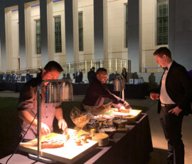 Two chefs serve food to a man at an outdoor buffet event in front of a large, well-lit building at night.