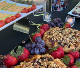 A table with fruit, cheese, and nut-topped pastries, with photos and candles, set outdoors under a tree.