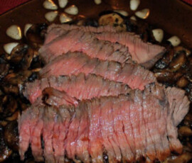 Sliced medium-rare roast beef served on a bed of sautéed mushrooms, garnished with garlic on a brown plate.