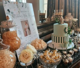 Dessert table with a green cake, candy jars, chocolates, and a baby shower welcome sign displayed by a window.