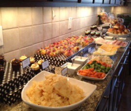 A kitchen counter with chips, cupcakes, fruit skewers, vegetables, cheese, bread, and sandwich fixings arranged for a buffet.