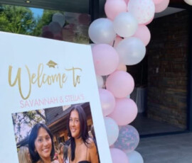 A graduation party welcome sign with a photo, pink balloon arch, and a wooden door in the background.