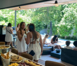 Women in white dresses gather inside and outdoors by a pool; a table is set with fruit, vegetables, and snacks.