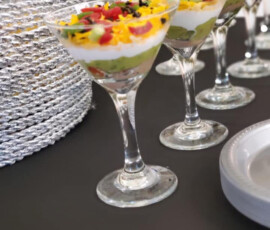 Several layered dip appetizers are served in martini glasses on a black table next to a stack of silver placemats.