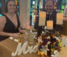 Two people in formal attire sit at a decorated table with candles and flowers in front of stained glass windows.