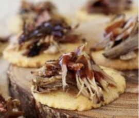 Screenshot Shredded meat with sauce served on round crackers, arranged on a wooden board.