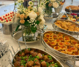 Buffet table with trays of meatballs, pastries, and other appetizers, decorated with fresh white and beige flowers.
