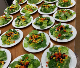 Rows of white plates with green salads topped with corn, black beans, and diced bell peppers on a wooden table.