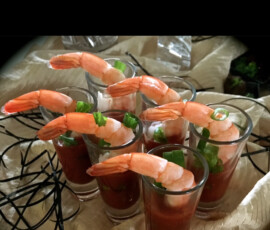 Six shrimp cocktail appetizers in shot glasses with cocktail sauce and green garnish, arranged on a decorative surface.