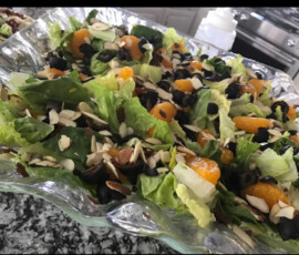 Salad with romaine lettuce, mandarin oranges, black olives, and sliced almonds in a clear glass bowl on a countertop.