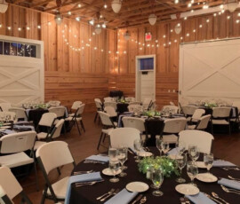 Rustic banquet hall with round tables set for dinner, string lights overhead, and wood-paneled walls.