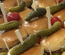 Mini sandwiches topped with pickles and skewered with wooden sticks, garnished with cherry tomatoes on a tray.